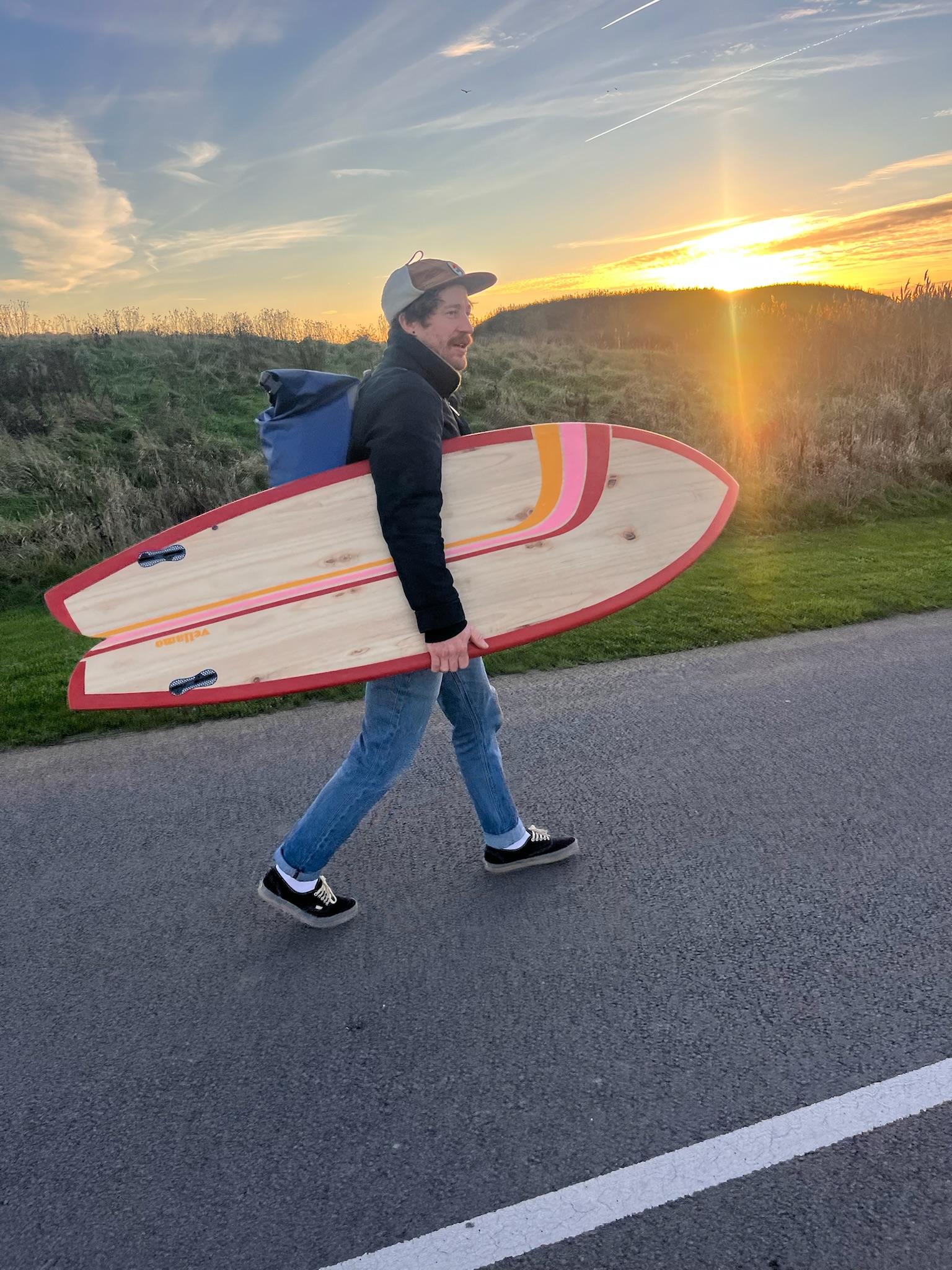 A surfer carrying a timber twin-fin down to the water at last light